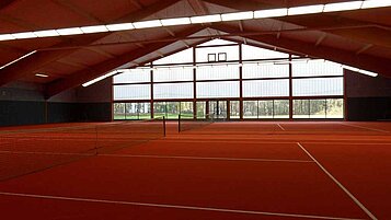 Innenansicht der modernen Tennishalle im Familienhotel Sonnenhügel mit hell erleuchteten, roten Tennisplätzen und großen Fenstern mit Ausblick auf die grüne Landschaft.