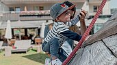 Ein Vater hält sein Baby fest, während dieses versucht auf dem Outdoor-Spielplatz eine Kletterwand hinauf zu klettern im Familienhotel Gorfion in Liechtenstein.