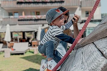 Ein Vater hält sein Baby fest, während dieses versucht auf dem Outdoor-Spielplatz eine Kletterwand hinauf zu klettern im Familienhotel Gorfion in Liechtenstein.