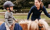 Kind sitzt auf dem Reitplatz des Familienhotels Landhaus Averbeck in der Lüneburger Heide auf einem Pony und wird von einer Reitlehrerin geführt.