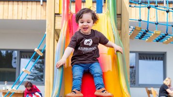 Kleinkind rutscht im Sommerurlaub in Kärnten eine bunte Rutsche auf dem Outdoor-Spielplatz des Familienhotels Petschnighof hinunter.