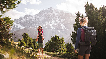 Vater trägt seine Tochter beim Wandern in Südtirol auf den Schultern. Die Mutter geht auf die beiden zu.