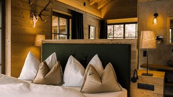 Gemütliches Schlafzimmer im Alpura Retreat mit Holzwänden, großen Fenstern, Hirschgeweih an der Wand und stilvoll dekoriertem Bett.