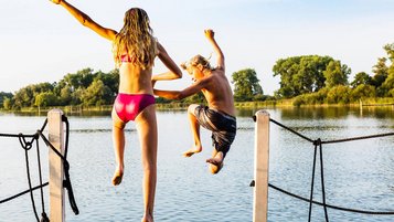 Zwei jugendliche Kinder springen vom Steg in einen See und heben vor Freude die Hände in die Höhe.