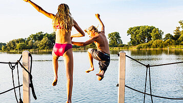 Zwei jugendliche Kinder springen vom Steg in einen See und heben vor Freude die Hände in die Höhe.