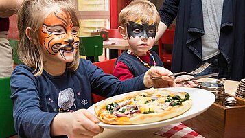 Zwei Kinder mit Gesichtsbemalung greifen begeistert nach einer Pizza in einem bunten Kinderrestaurant, was die familienfreundlichen Aktivitäten und das kindgerechte Essen im Familienhotel Deichkrone an der Nordsee unterstreicht.