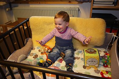 Ein glückliches Kleinkind sitzt mit verschiedenen Spielsachen im Gitterbett im Spa- & Familien-Resort Krone im Allgäu.