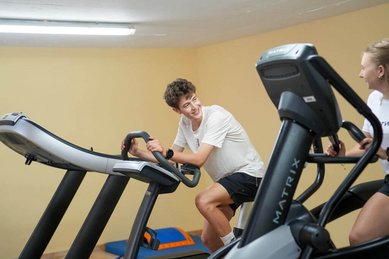 Zwei Jugendliche trainieren lachend im Fitnessraum des Familienhotel Sonne Bezau auf Laufband und Crosstrainer.
