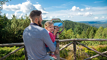 Vater und Tochter gucken gemeinsam durch ein Fernrohr beim Wandern im Thüringer Wald und erkunden die schöne Landschaft.