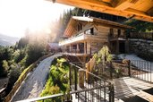 Großzügiges Holzchalet im Alpura Retreat in Hanglage, umgeben von Wald und Natur, mit Sonnenschein und Blick auf eine Schotterstraße und Terrassen.