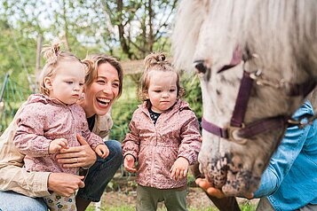 Mutter steht mit ihren zwei Kleinkindern vor einem Pony das gerade gefüttert wird im Familienhotel Das Hopfgarten in Tirol.