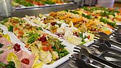 Nahaufnahme eines appetitlichen Buffets im Familienhotel Deichkrone mit einer Auswahl an Salaten, Wurst, Käse und frischem Obst, arrangiert auf weißen Servierplatten, bereit zum Genuss für die Gäste.