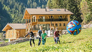 Vier Kinder spielen auf der grünen Wiese vor dem Familienhotel Huber in Südtirol bei schönem Wetter mit dem Ball.