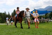 Ein Mädchen reitet auf einem Pony, begleitet von ihrer Familie auf einer Wiese des Familienhotels Ramsi in Kärnten