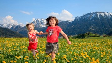 Zwei Kinder laufen über eine Wiese voller Löwenzahn vor der Alpenkulisse im Allgäu.