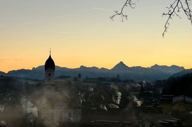 Sonnenaufgang über Nesselwang mit Kirchturm und Bergkulisse nahe dem Familienhotel Bavaria.