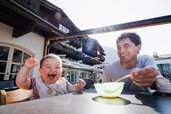 Ein lachendes Baby sitzt mit einem Erwachsenen an einem Tisch auf der Sonnenterrasse des Familienhotels Tirolerhof beim Füttern.