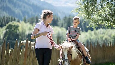 Ein kleienr Junge sitzt auf dem Pony und wird von einer Betreuerin geführt im Familienhotel Alphotel Tyrol Wellness & Family Resort in Südtirol.