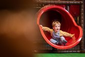 Ein Kleinkind saust lachend die rote Rutsche im Indoor-Spielbereich des Familienhotels Elldus im Erzgebirge hinunter.