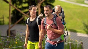 Familie mit Kleinkind in der Rückentrage beim Wandern vor dem Familienhotel Das Bayrischzell in den Bergen.