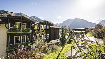 Sonnendurchfluteter Garten mit Spielplatz vor einem traditionellen alpinen Holzhaus, umgeben von einem zauberhaften Bergpanorama, ideal für Familienurlaub in natürlicher Umgebung.