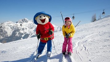 Ein glückliches Kind fährt im Familienhotel Ramsi in Kärnten mit dem Maskottchen auf der Skipiste, umgeben von einer beeindruckenden Bergkulisse