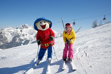 Ein glückliches Kind fährt im Familienhotel Ramsi in Kärnten mit dem Maskottchen auf der Skipiste, umgeben von einer beeindruckenden Bergkulisse