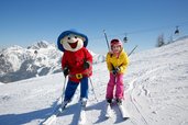 Ein glückliches Kind fährt im Familienhotel Ramsi in Kärnten mit dem Maskottchen auf der Skipiste, umgeben von einer beeindruckenden Bergkulisse