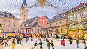 Mitten in der Stadt steht eine Eisbahn voll mit Menschen. Die Stadt in Kaernten ist schön weihnachtlich geschmückt.