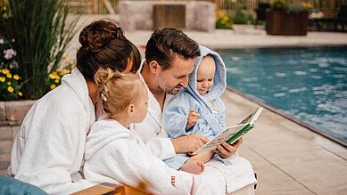 Familie mit zwei Kindern sitzt im Bademantel am Außenpool des Familienhotels Landhaus Averbeck in der Lüneburger Heide. Der Vater liest ein Buch vor.