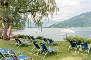 Mehrere leere Sonnenliegen unter Bäumen und einem weißen Sonnenschirm auf dem hoteleigenen Badestrand des Familienhotels Amiamo am Zeller See, dahinter Bergpanorama.