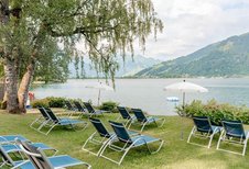Mehrere leere Sonnenliegen unter Bäumen und einem weißen Sonnenschirm auf dem hoteleigenen Badestrand des Familienhotels Amiamo am Zeller See, dahinter Bergpanorama.