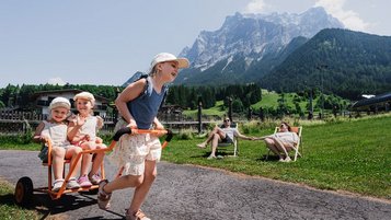 Drei Kinder spielen mit einem Rutschfahrzeug vor der Bergkulisse, während zwei Erwachsene entspannt auf Liegestühlen vor dem Familienhotel Tirolerhof sitzen.