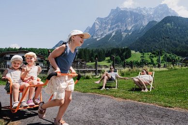 Drei Kinder spielen mit einem Rutschfahrzeug vor der Bergkulisse, während zwei Erwachsene entspannt auf Liegestühlen vor dem Familienhotel Tirolerhof sitzen.