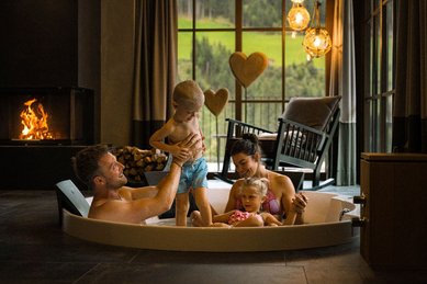 Familie mit zwei kleinen Kindern entspannt gemeinsam in einer Badewanne vor einem Kamin im Alpura Retreat, gemütliche Atmosphäre mit Blick ins Grüne.