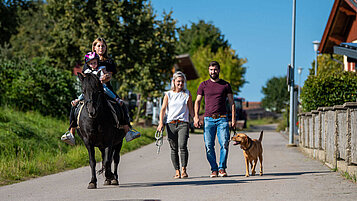 Familie bei einem Spaziergang mit Hund rund um das Familienhotel Der Böhmerwald. Das Kind sitzt gemeinsam mit einer Betreuerin auf einem Pferd.