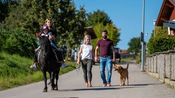 Familie bei einem Spaziergang mit Hund rund um das Familienhotel Der Böhmerwald. Das Kind sitzt gemeinsam mit einer Betreuerin auf einem Pferd.