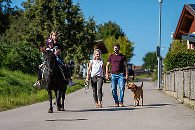 Familie bei einem Spaziergang mit Hund rund um das Familienhotel Der Böhmerwald. Das Kind sitzt gemeinsam mit einer Betreuerin auf einem Pferd.