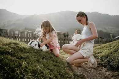 Kinder sitzen auf einer Wiese im Familienhotel Mia Alpina in Tirol und streicheln Hasen vor Bergkulisse.