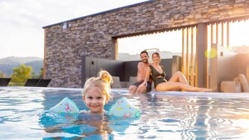 Mädchen schwimmt mit Schwimmflügeln im Außenpool des Familienhotels Schreinerhof, die Eltern sitzen am Beckenrand und passen auf,.