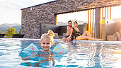 Mädchen schwimmt mit Schwimmflügeln im Außenpool des Familienhotels Schreinerhof, die Eltern sitzen am Beckenrand und passen auf,.