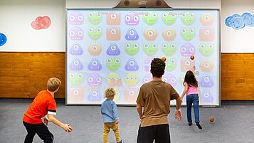Kinder spielen begeistert ein interaktives Ballspiel vor einer großen digitalen Spielwand im Spielzimmer des Hotels Feldberger Hof.