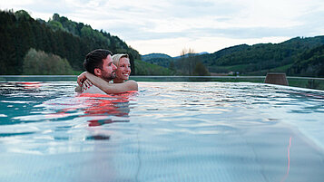 Eltern umarmen sich im Outdoor-Pool des Familienhotels und blicken in die Berglandschaft.