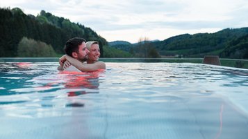 Ein Paar genießt den Abend in einem Infinity-Pool im Familienhotel mit Blick auf eine grüne Hügellandschaft