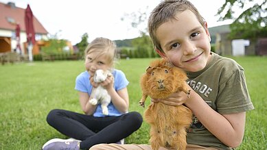 Ein Junge und ein Mädchen sitzen in der Wiese und kuscheln mit den Meerschweinchen des Familienhotels Rhön Feeling.