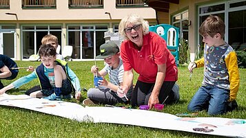 Kinder bemalen gemeinsam mit einer Kinderbetreuerin ein großes Plakat draußen in der Wiese des Familienhotels Lärchenhof in Tirol.
