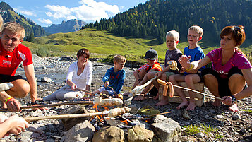 Familie beim Stockbrot grillen im Familienurlaub in Vorarlberg, umgeben von idyllischer Berglandschaft und Bachläufen.