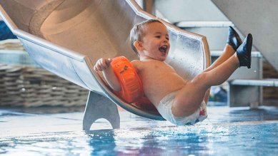 Kleines Kind mit Schwimmflügeln rutscht lachend ins Wasser im Hallenbad des Familienhotels Amiamo.