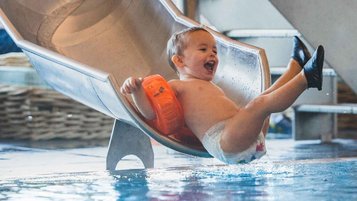 Kleines Kind mit Schwimmflügeln rutscht lachend ins Wasser im Hallenbad des Familienhotels Amiamo.