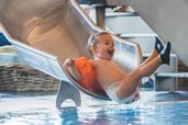 Kleines Kind mit Schwimmflügeln rutscht lachend ins Wasser im Hallenbad des Familienhotels Amiamo.